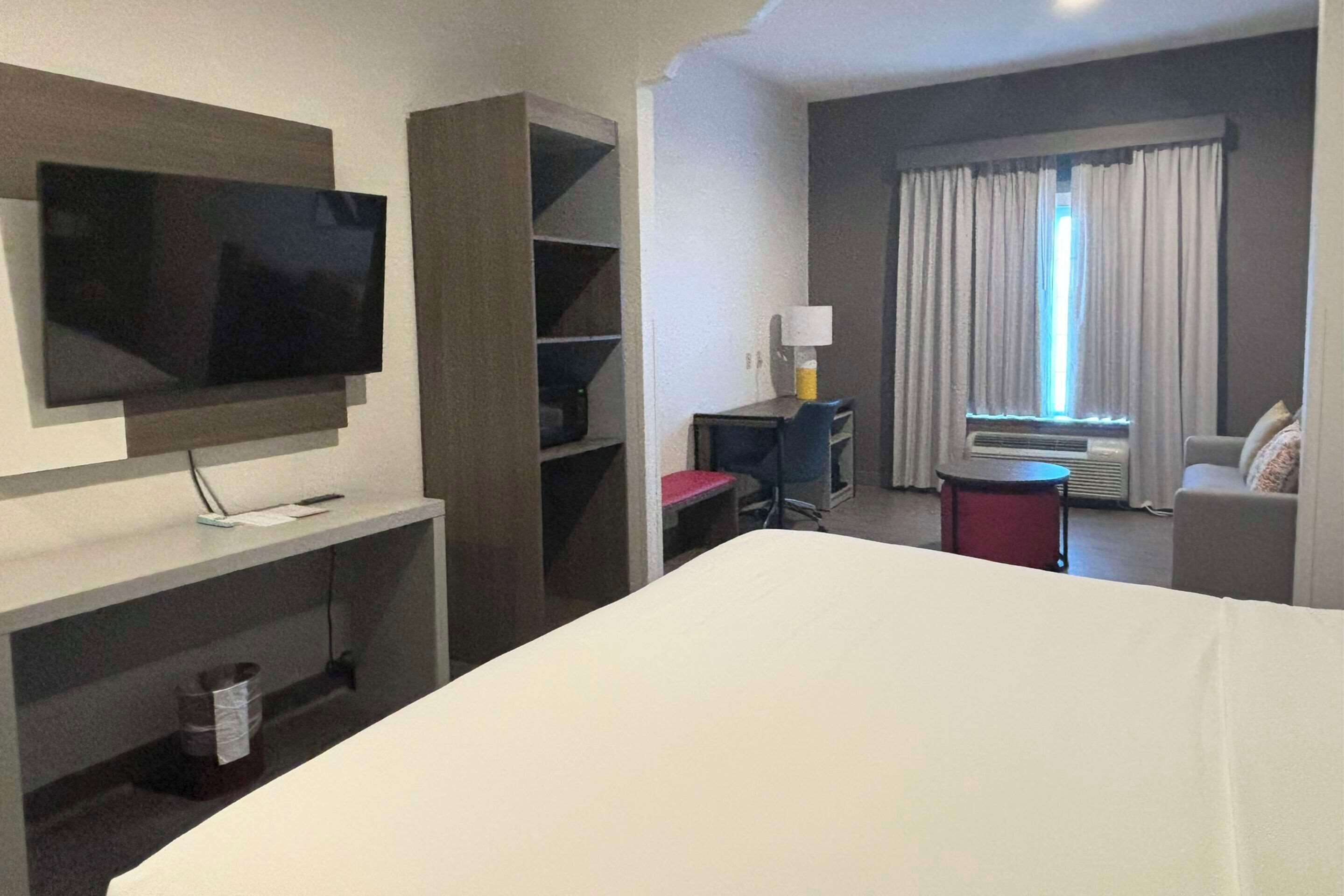 A hotel room with a bed, a TV mounted on the wall, a desk with a chair, a lamp, and a window with curtains.