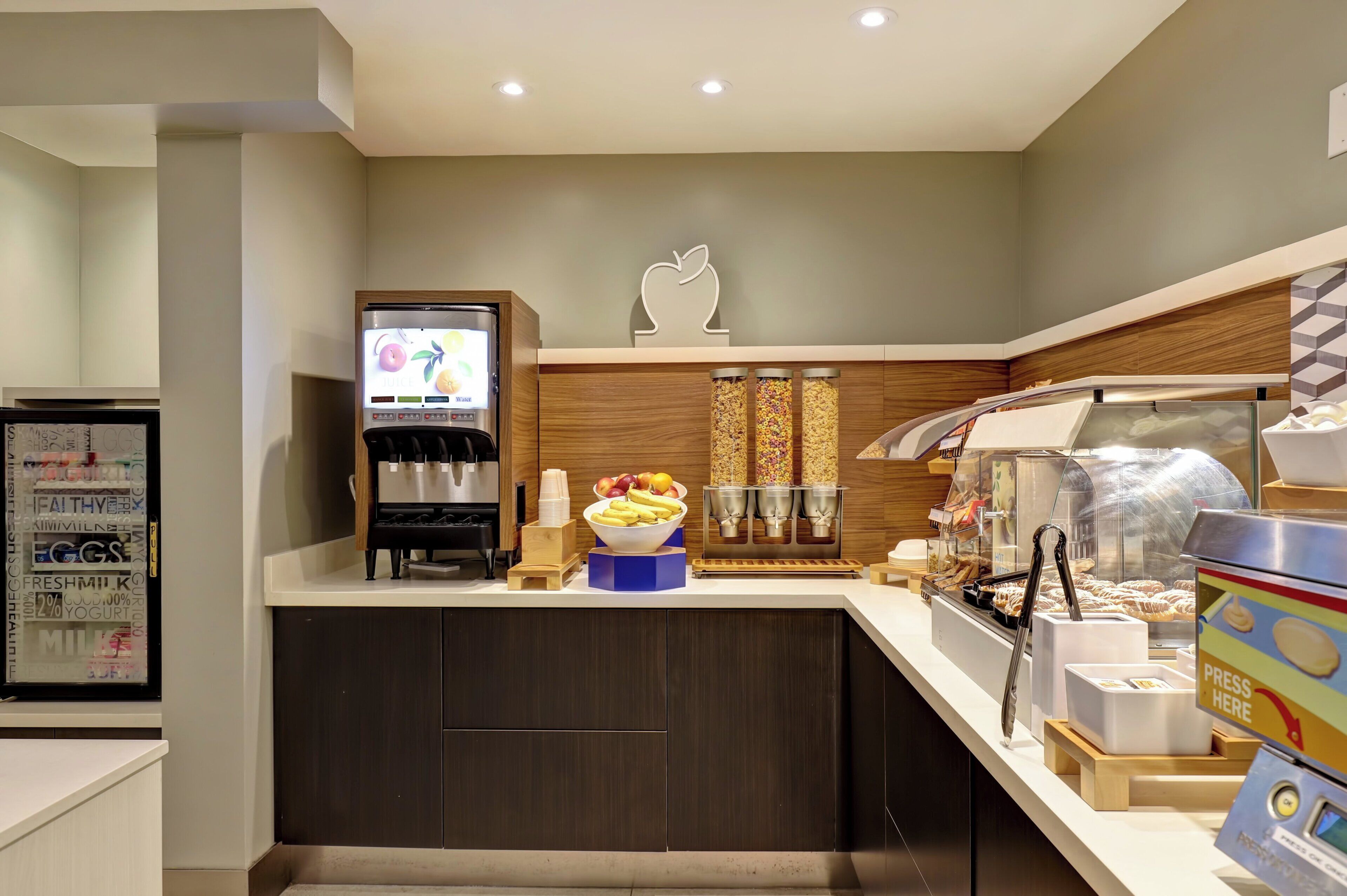 A self-service breakfast area with a coffee machine, fruit bowl, and a display case with pastries.