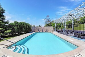 A rectangular swimming pool with a stone wall and a covered seating area with chairs.