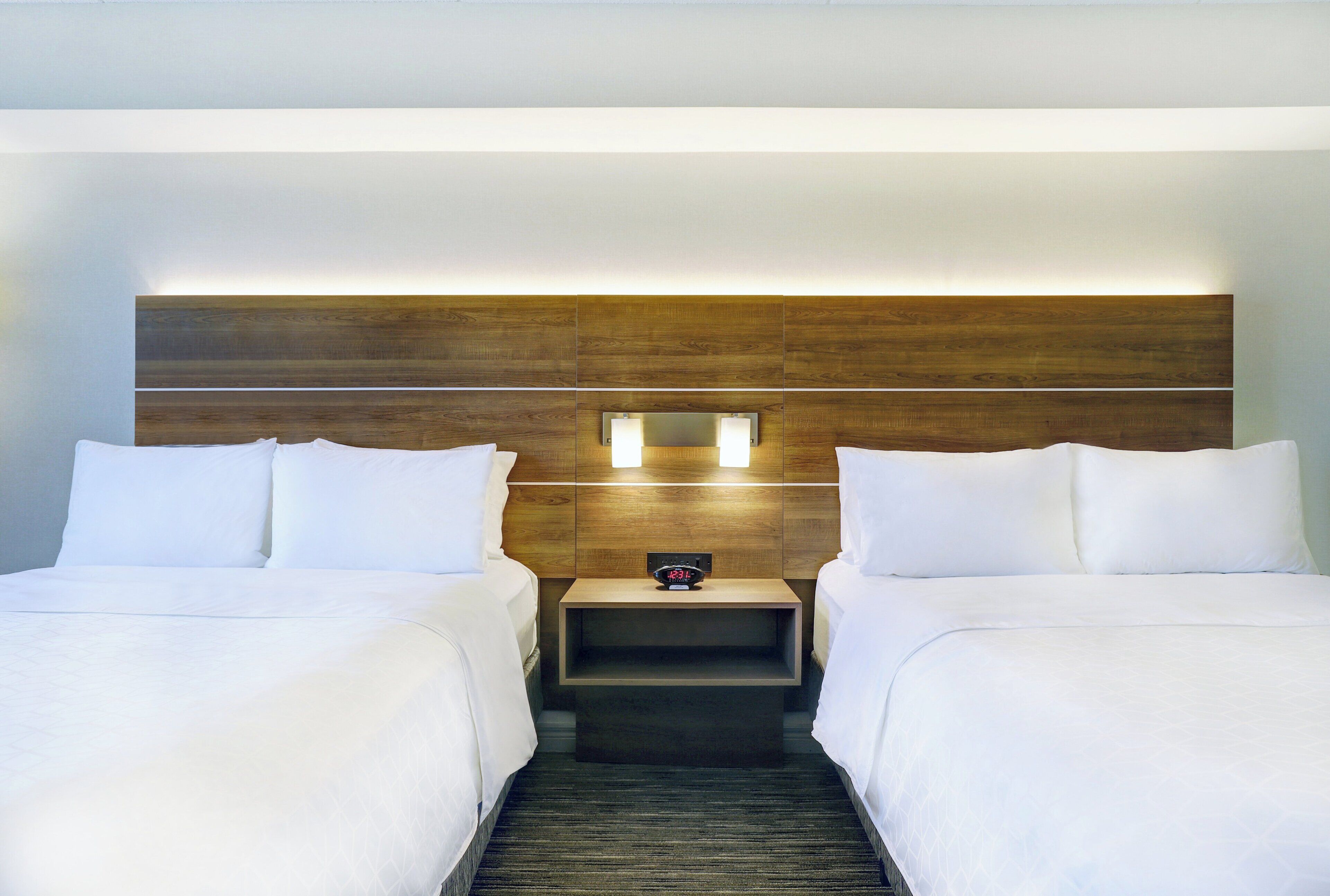 A hotel room with two beds, a wooden headboard, a nightstand with a clock, and a carpeted floor.
