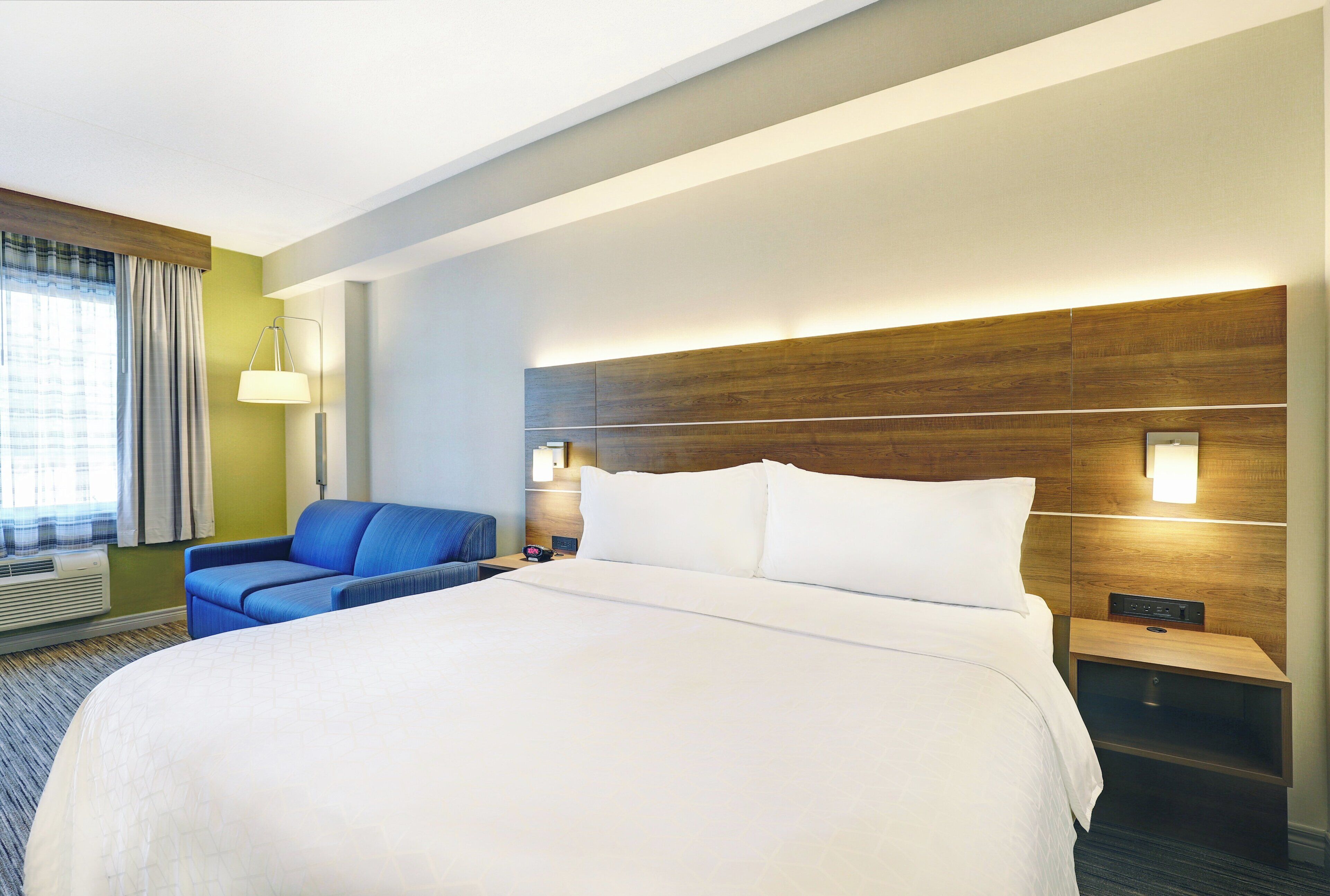 A hotel room with a large bed, a blue armchair, a nightstand with a lamp, and a wall-mounted TV.