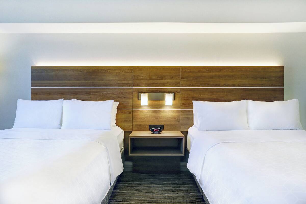 A hotel room with two beds, a wooden headboard, a nightstand with a clock, and a small shelf.