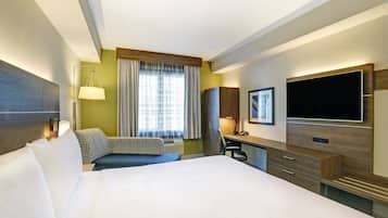 A hotel room with a large bed, a desk with a flat-screen TV, a chair, and a window with curtains.