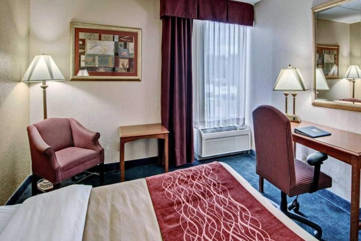Una habitación de hotel con una cama, dos sillas, un escritorio, una lámpara, un marco de fotos, una ventana con cortinas y un equipo de aire acondicionado.
