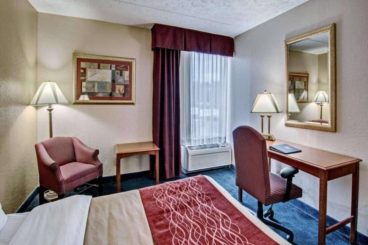 Habitación de hotel con cama, dos sillones, escritorio, dos lámparas, ventana con cortinas e imagen en el marco en la pared.