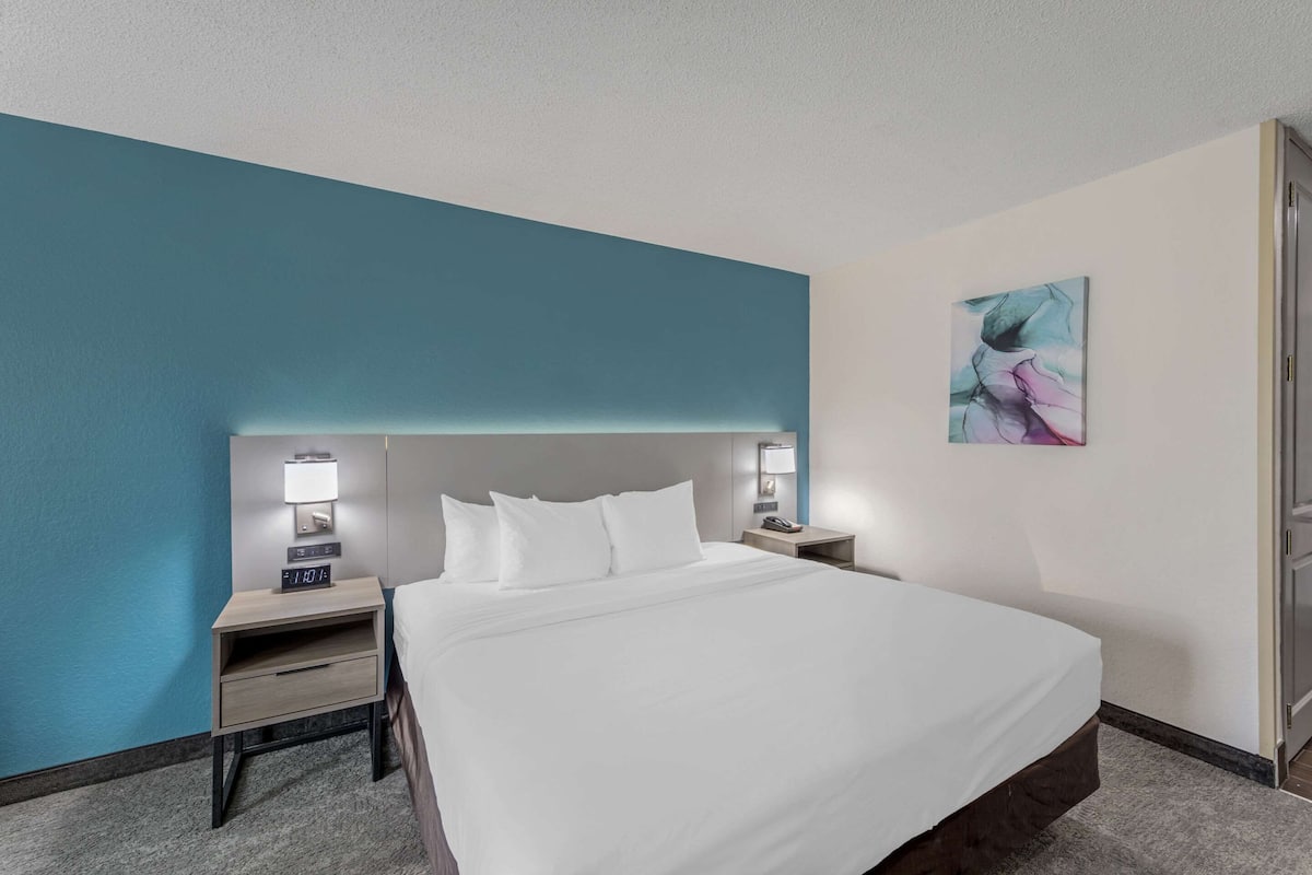 A hotel room with a large bed, two bedside tables with lamps, a wall-mounted artwork, and a door to the right.