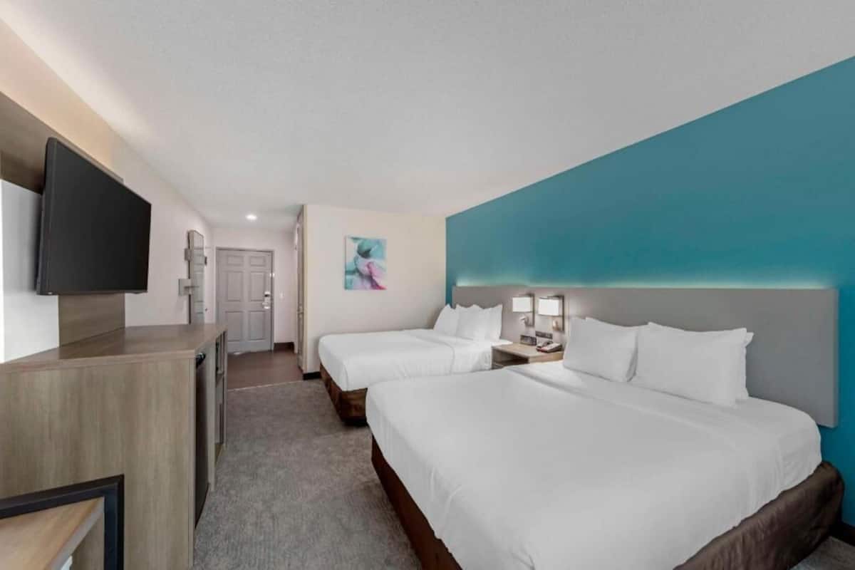 A hotel room with two beds, a desk, a flat-screen TV, a painting, and a bathroom in the background.