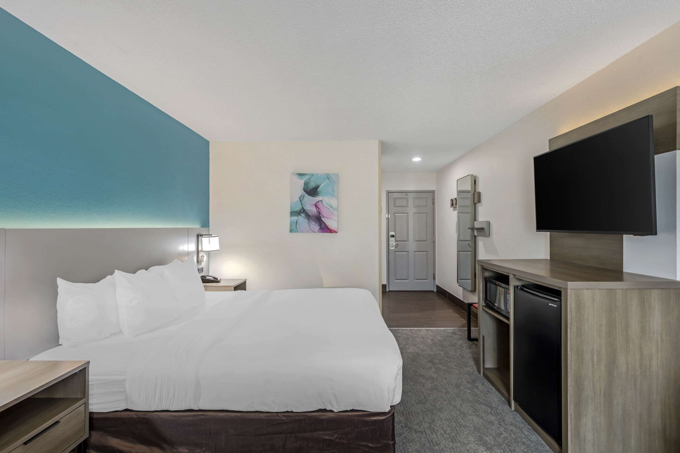 Habitación de hotel con cama, mesita de noche, lámpara, obra de arte, televisor y kitchenette.