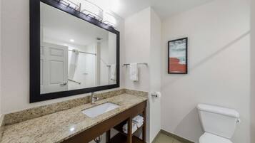 Un baño con lavamanos de encimera de granito, un espejo grande, un inodoro blanco y un cuadro en la pared.