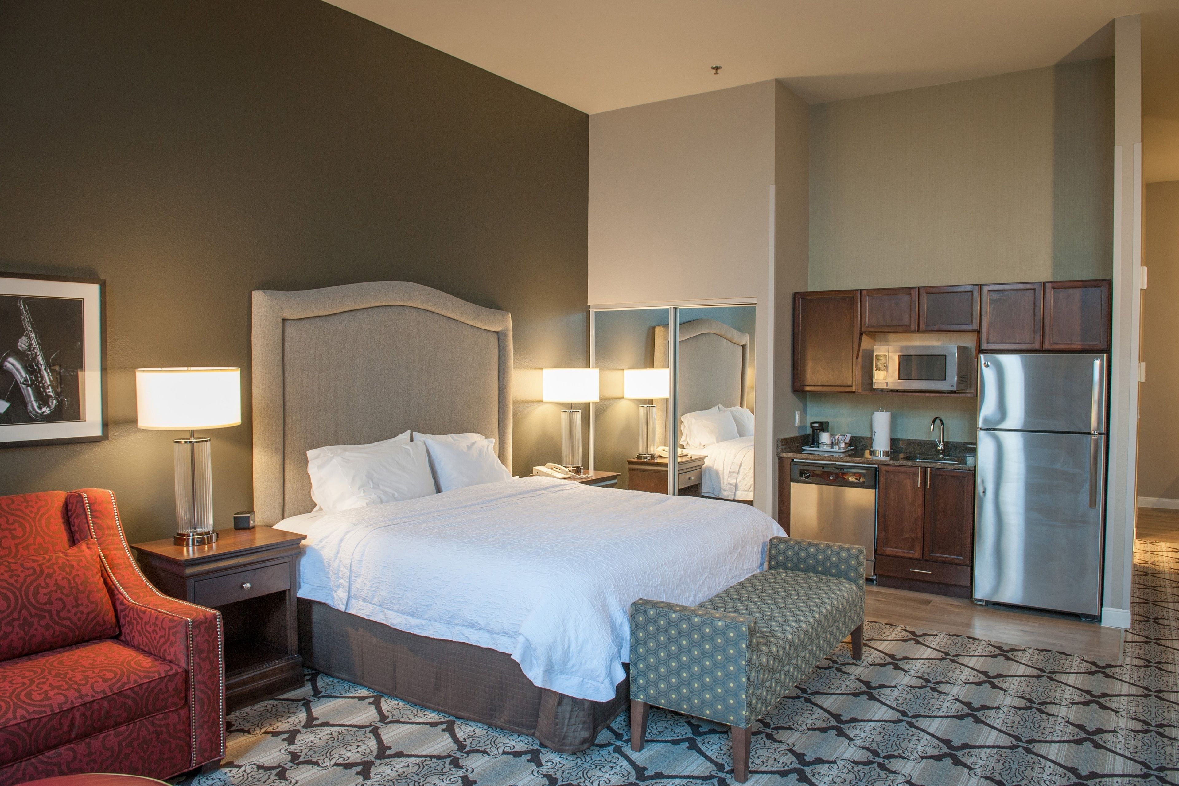 A hotel room with a bed, bedside tables, a desk, a chair, a kitchenette, and a patterned floor.
