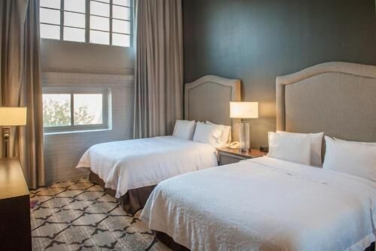 A hotel room with two beds, a window with curtains, a patterned floor, and a nightstand with a lamp.