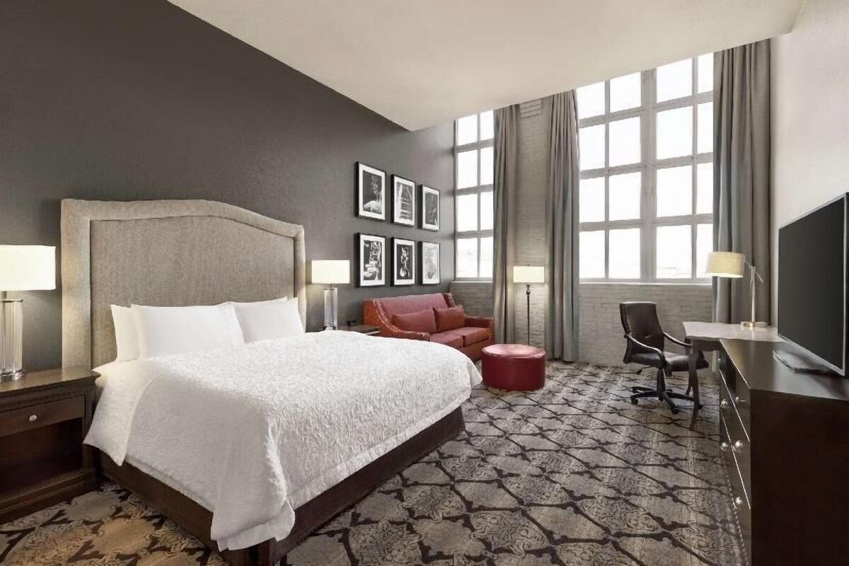A hotel room with a large bed, a desk, a chair, a red ottoman, and a TV.