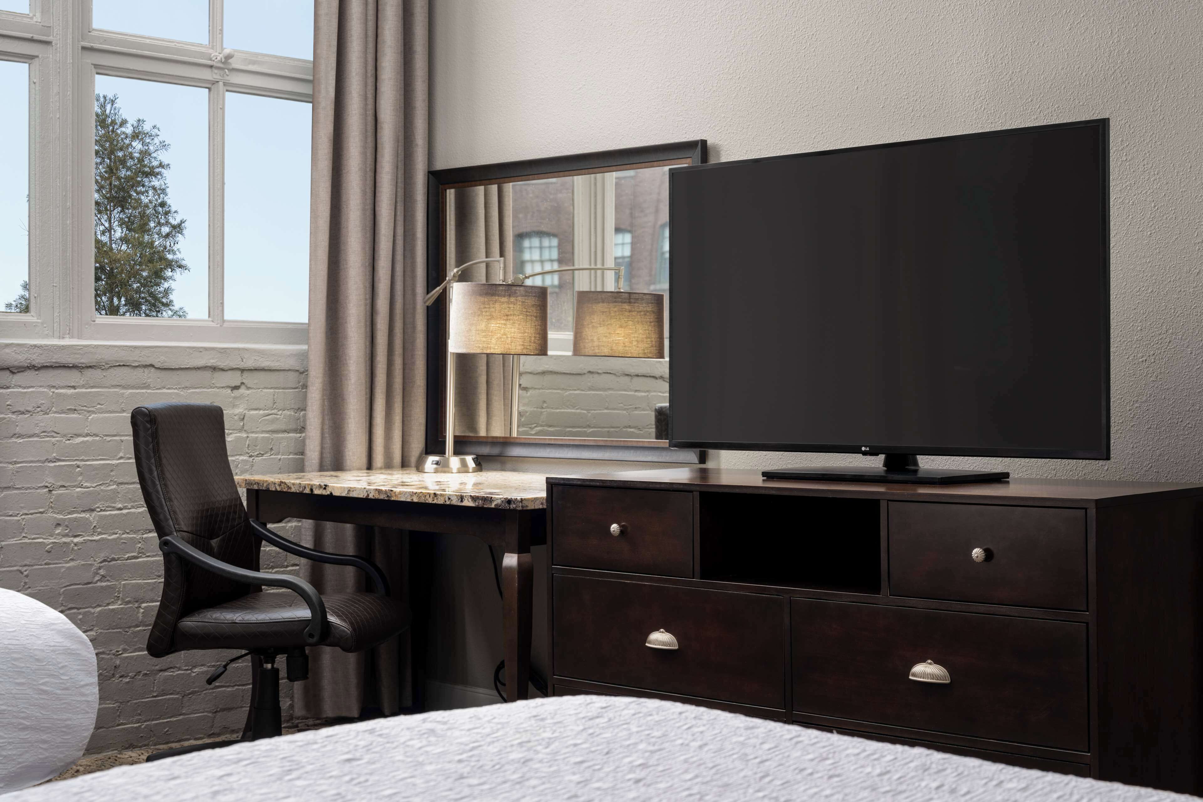 A hotel room with a bed, a desk with a chair, a TV, and a lamp.