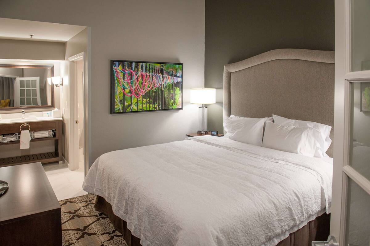 A hotel room with a large bed, a bedside table, a desk, a chair, a lamp, a mirror, and a painting on the wall.
