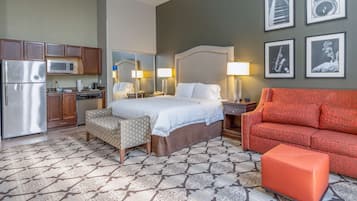 A hotel room with a bed, a sofa, a small table, a nightstand, a mirror, a kitchen area with a refrigerator and microwave, and a patterned floor.