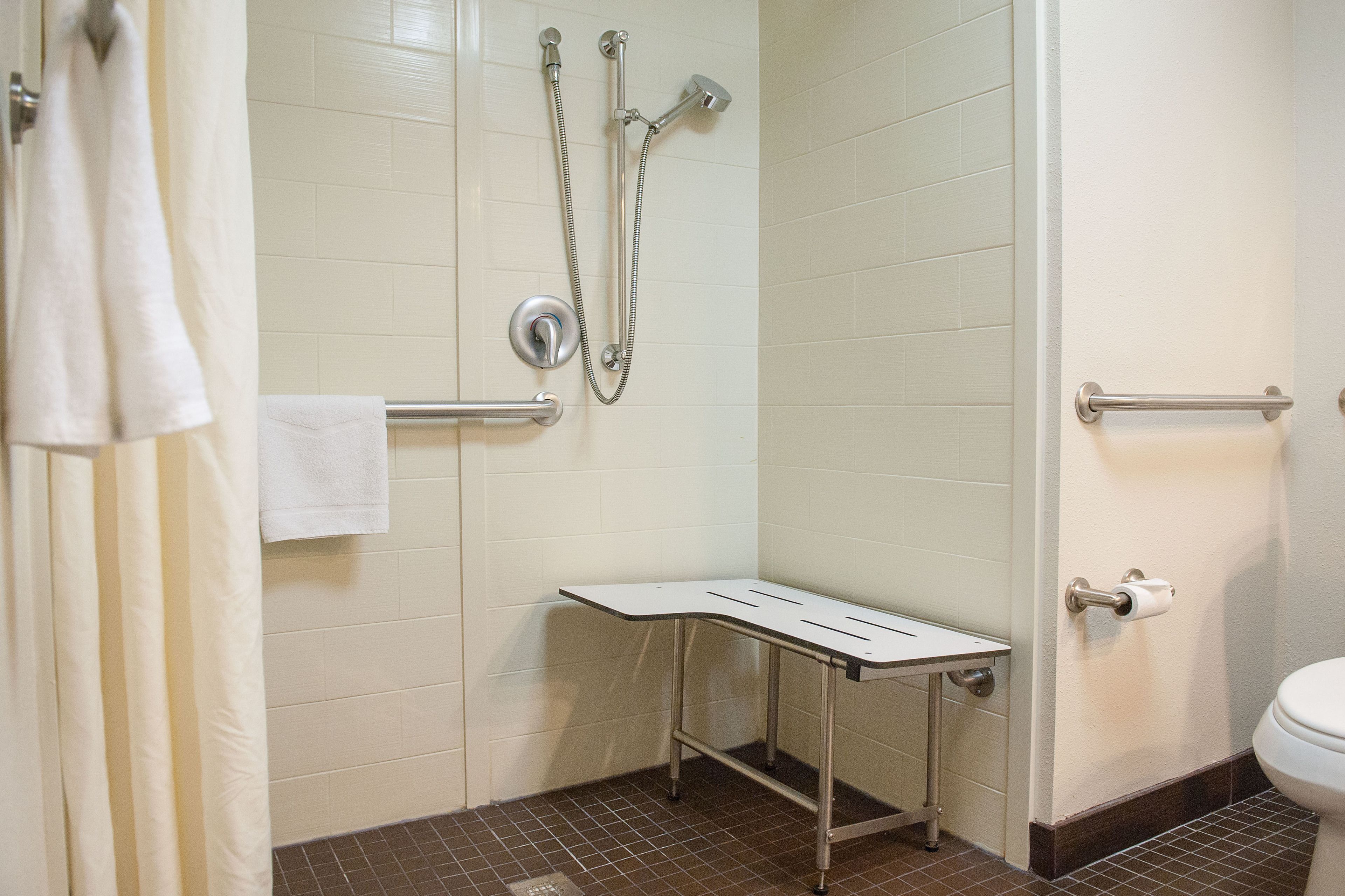 A bathroom with white tiled walls, a shower with a handheld showerhead, a toilet with a paper towel holder, and a towel rack.