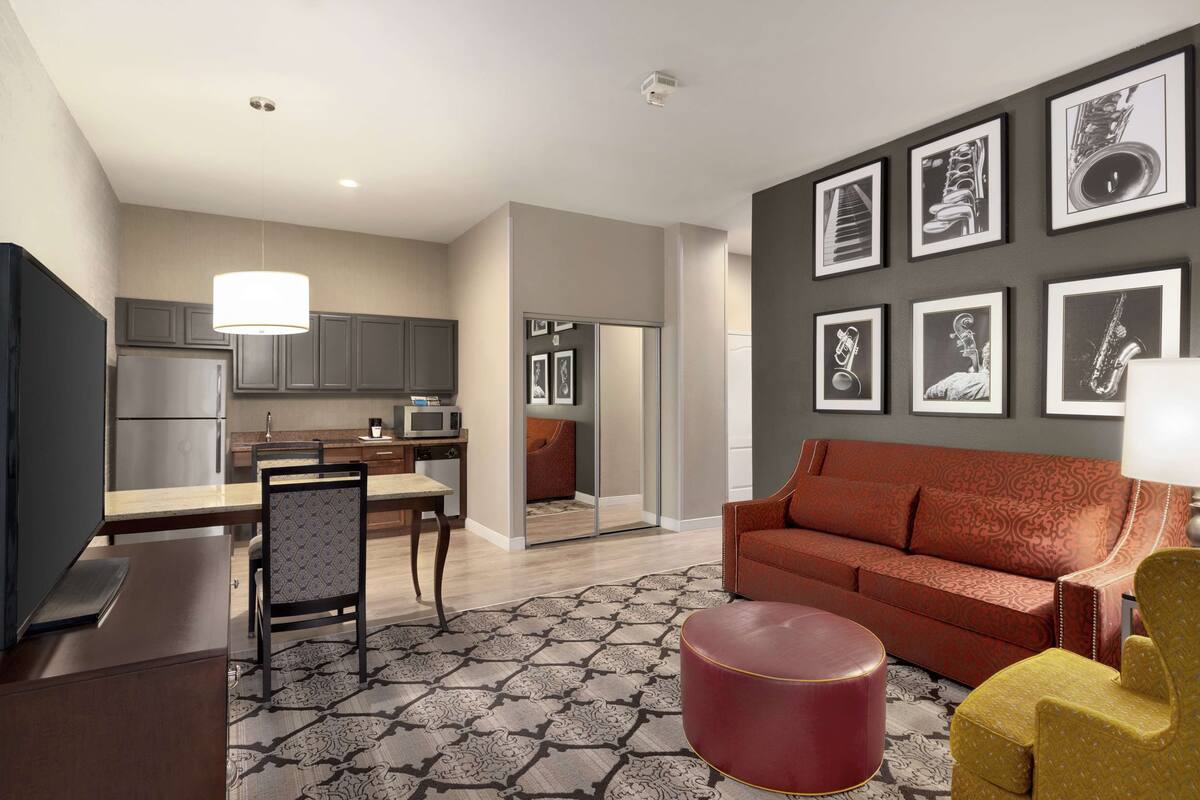 A modern living room with a sofa, a red ottoman, and a yellow armchair. There is a dining area with a table and chairs, and a kitchen with cabinets and a refrigerator.