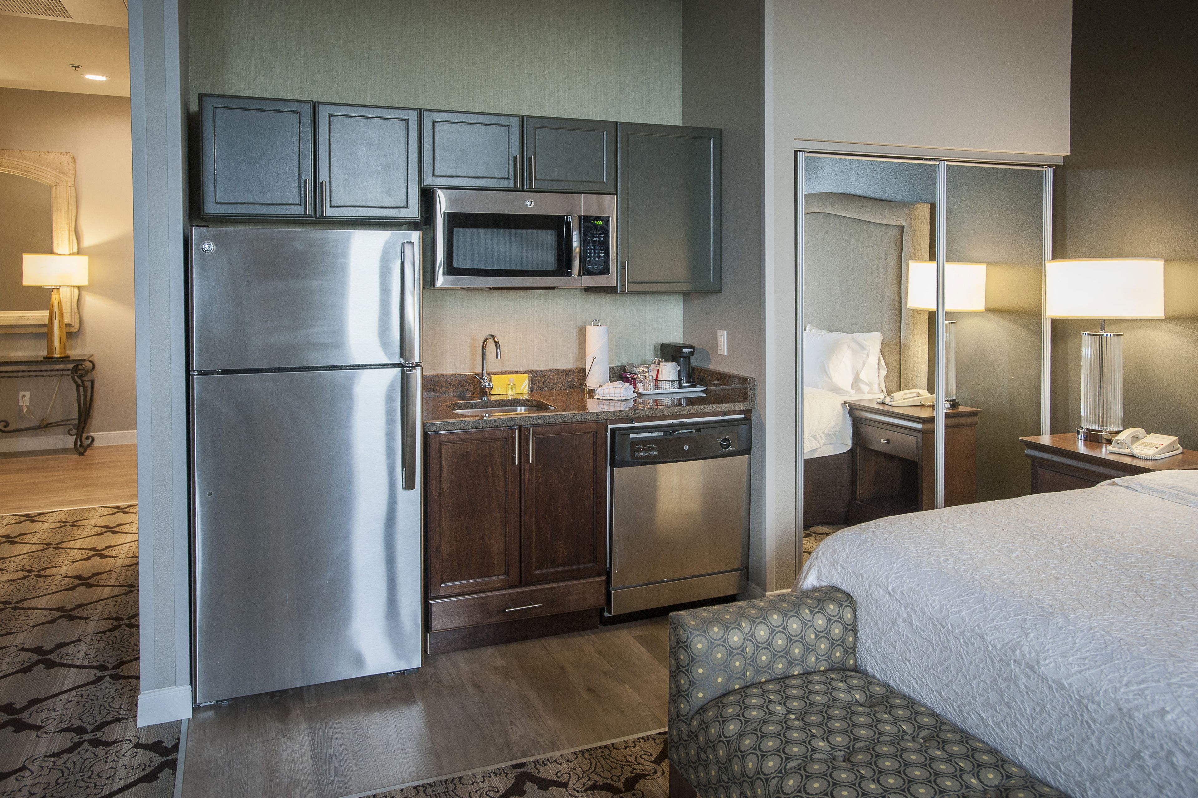 A compact hotel room with a kitchenette including a refrigerator, microwave, and dishwasher. There is a bed with a patterned headboard and a bedside table with a lamp.