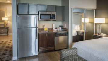 A compact hotel room with a kitchenette including a refrigerator, microwave, and dishwasher. There is a bed with a patterned headboard and a bedside table with a lamp.