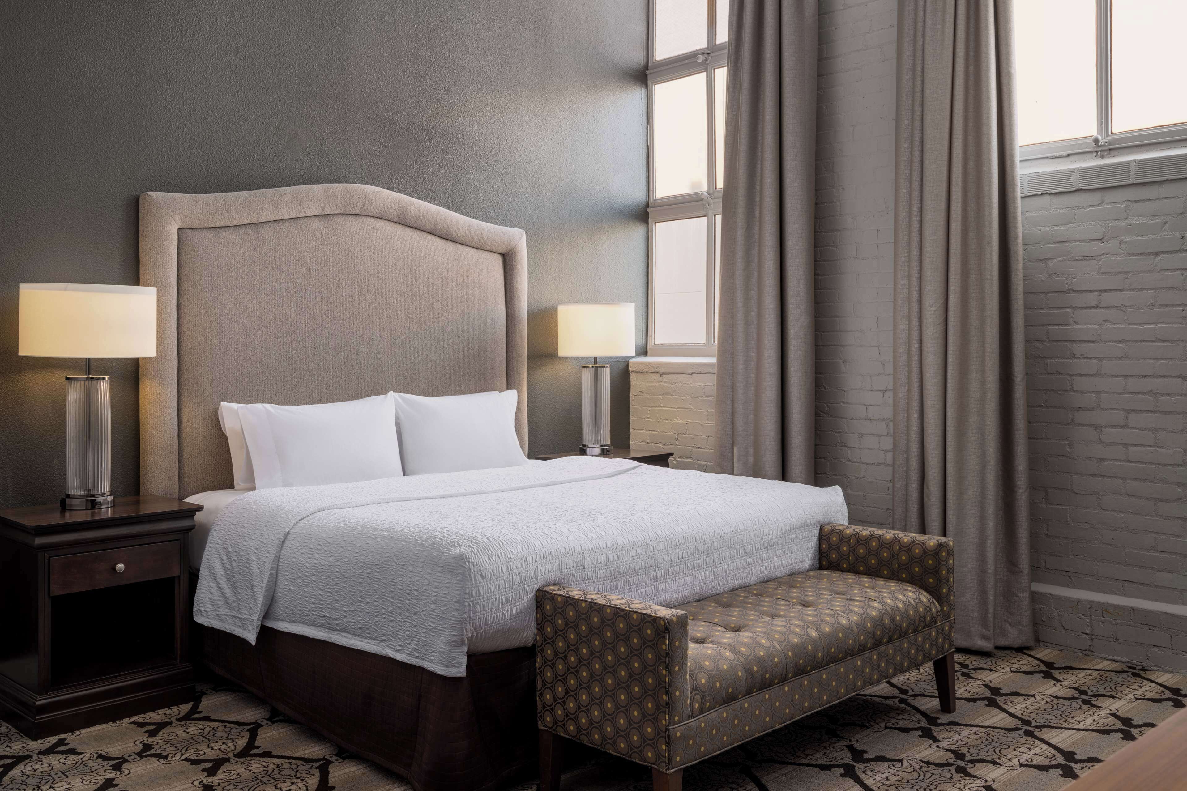 A neatly made bed with a headboard, two bedside lamps, a patterned armchair, and a window with curtains.