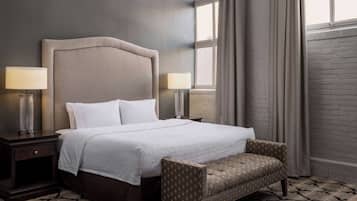 A neatly made bed with a headboard, two bedside lamps, a patterned armchair, and a window with curtains.