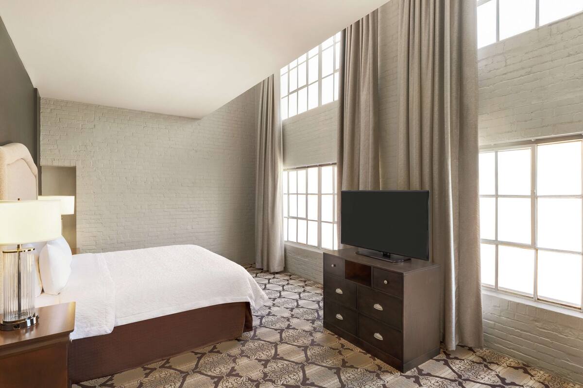 A hotel room with a bed, a TV on a stand, a lamp, and a large window with curtains.