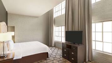 Quarto de hotel com cama, televisão sobre suporte, abajur e uma janela grande com cortinas.