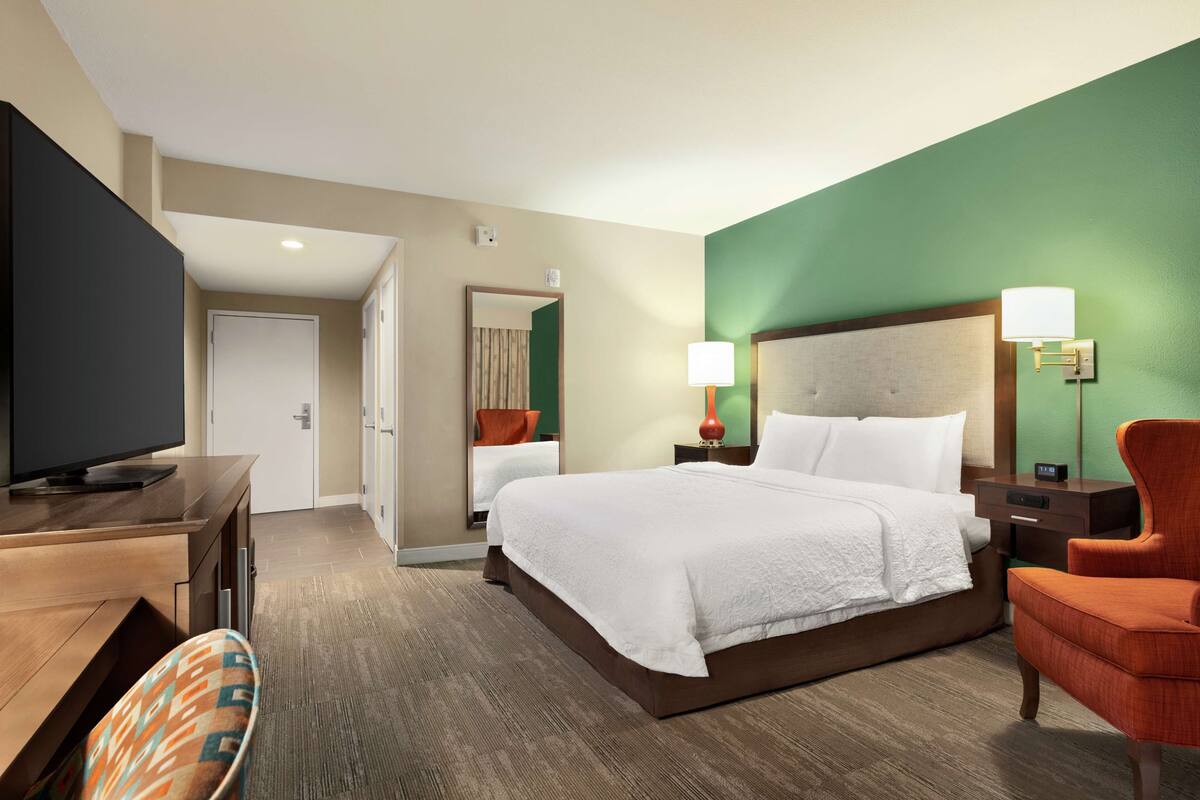 A hotel room with a large bed, a desk with a television, a chair, and a nightstand with lamps.