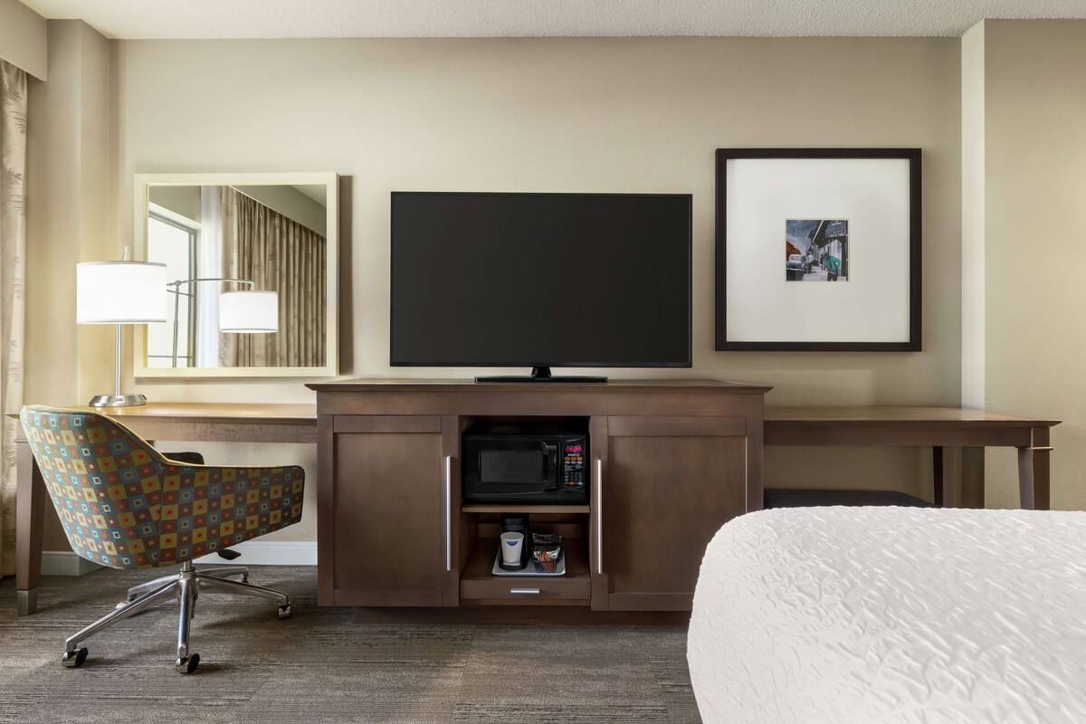 A hotel room with a bed, a desk with a flat-screen TV, a microwave, a chair, and a framed picture on the wall.