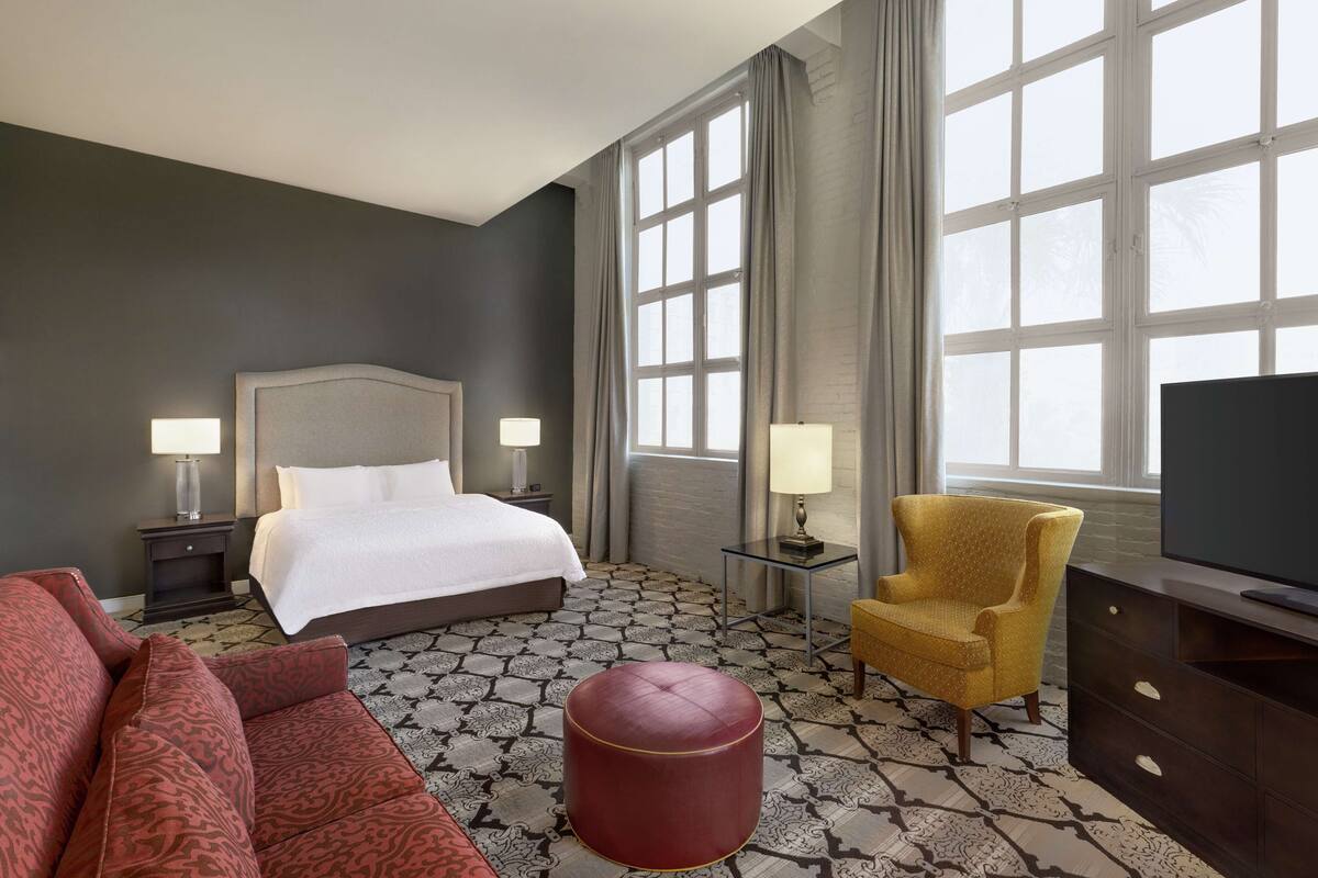 A hotel room with a bed, two bedside tables with lamps, a sofa, a chair, a television, and a patterned floor.
