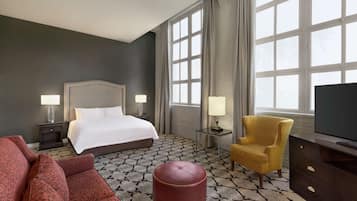 Habitación de hotel con cama, dos mesitas de noche con lámparas, sofá, sillón, televisor y suelo estampado.