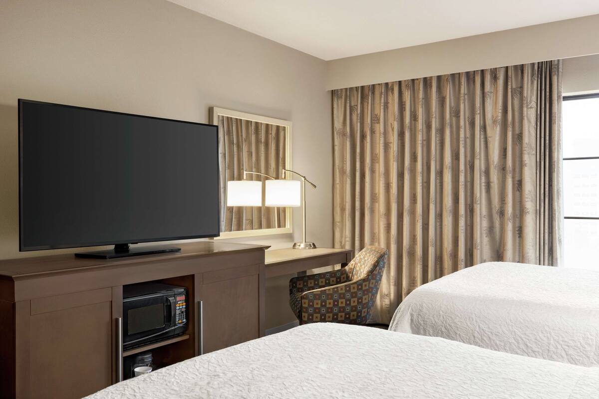 A hotel room with a flat-screen TV, a desk with a lamp, a microwave, two beds, and a chair.