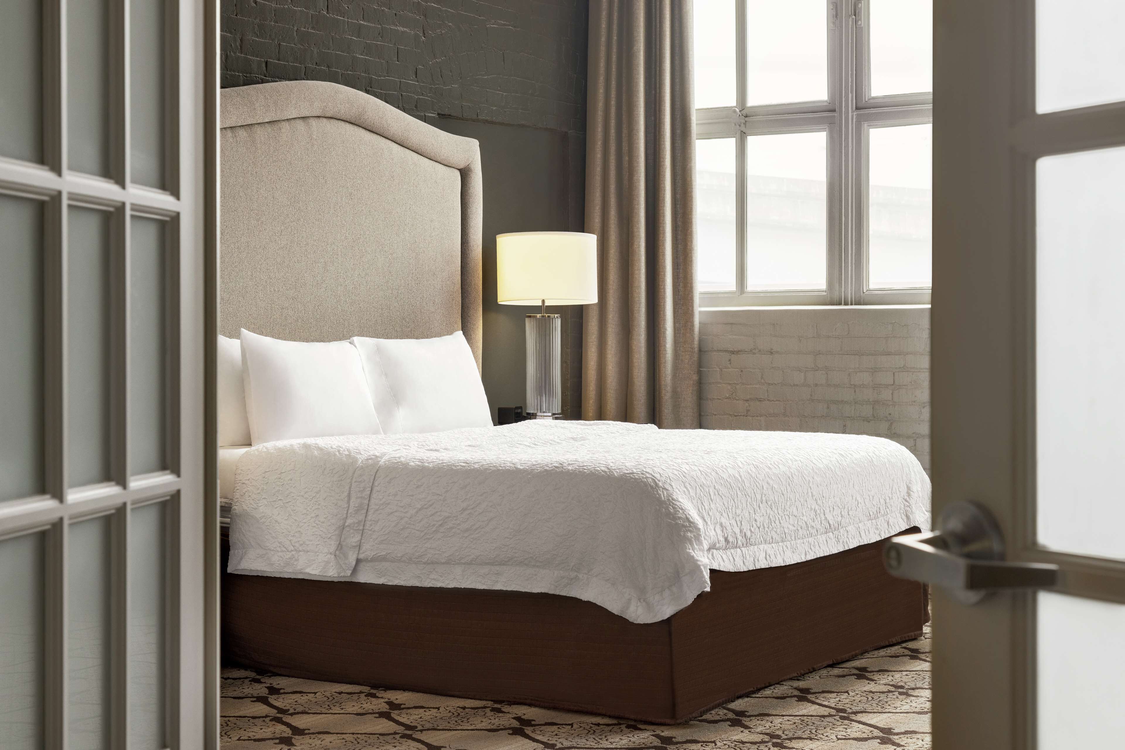 A neatly made bed with a headboard, a bedside lamp, and a view of a brick wall and window.