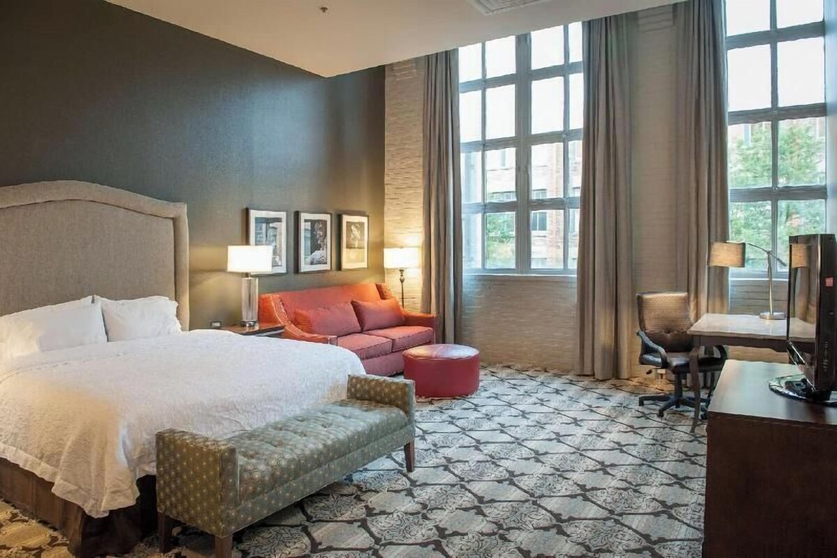 A hotel room with a bed, a red sofa, a desk, and a large window.