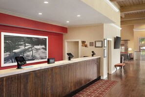 A hotel reception area with a wooden reception desk, a large framed picture on the wall, and a view into the dining area.