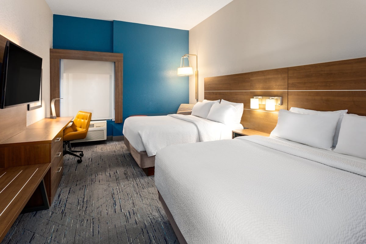 A hotel room with two beds, a desk, a chair, a TV, and a wall-mounted lamp.