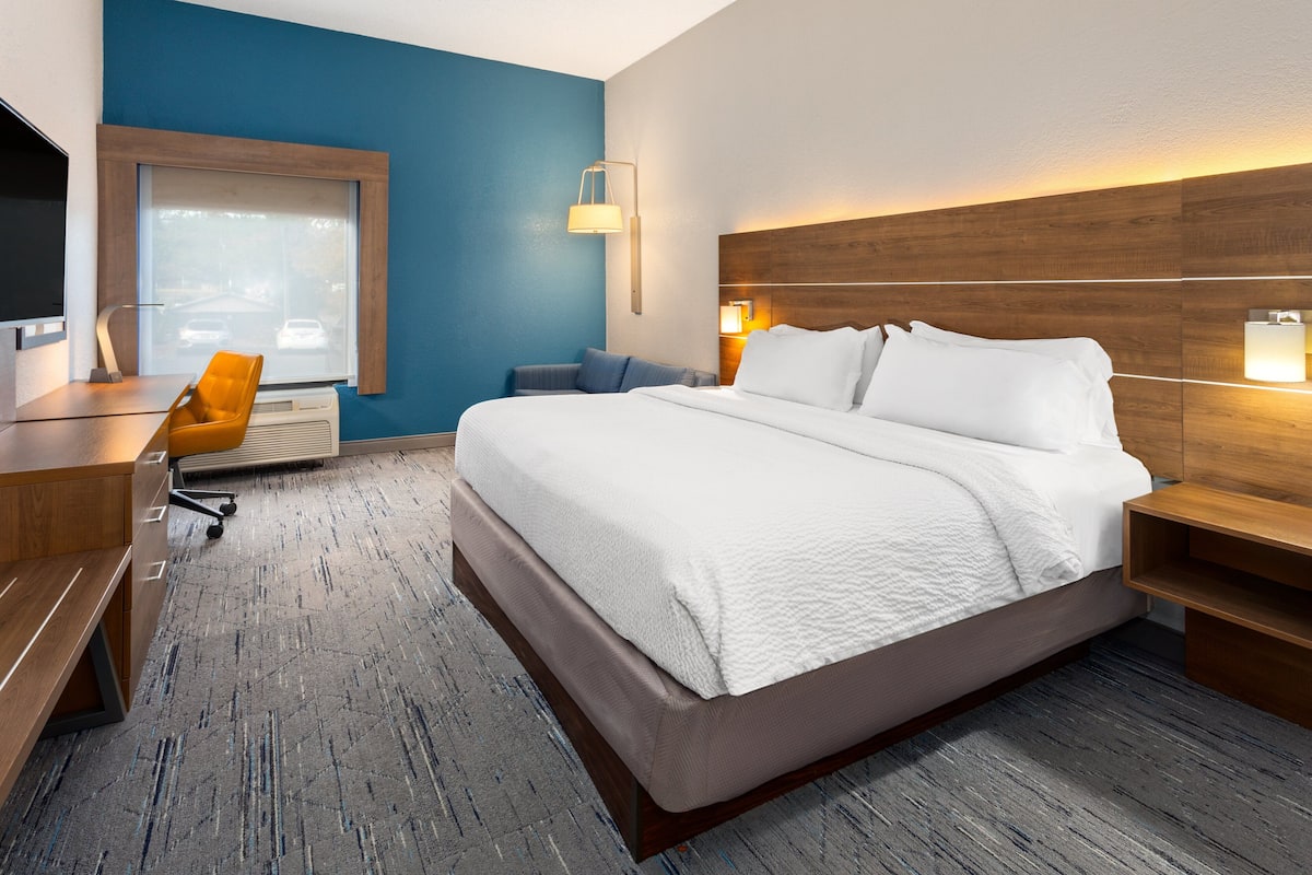 A hotel room with a large bed, a desk with a chair, a TV, and a window with a view of parked cars.