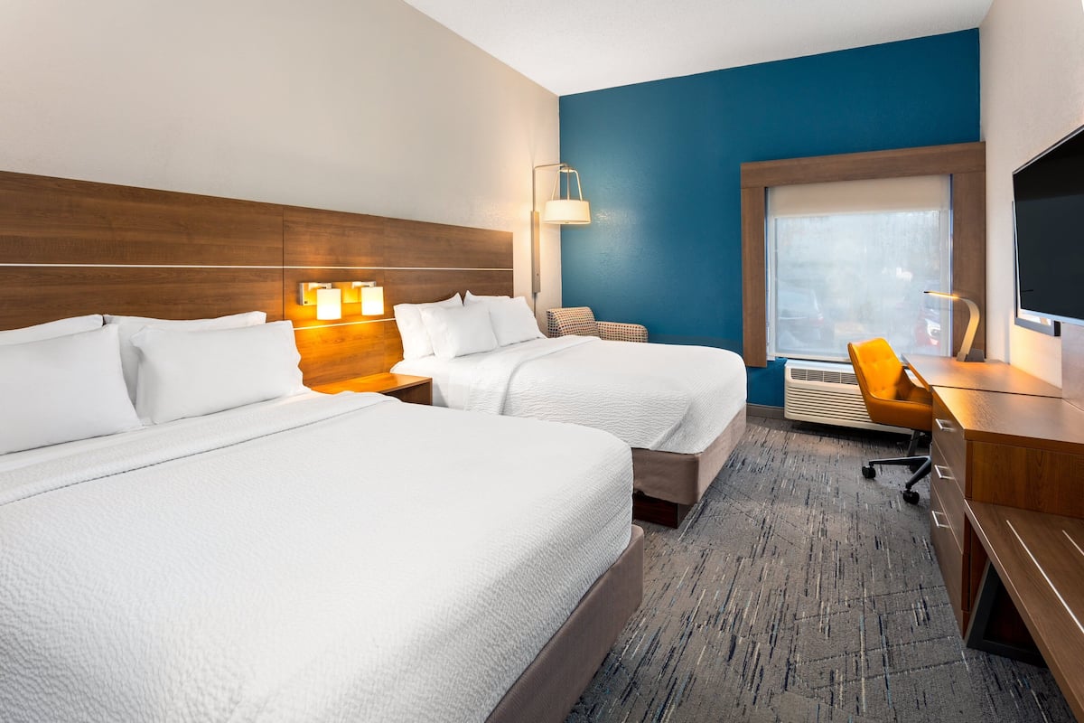 A hotel room with two beds, a desk, a chair, a TV, and a window with a view of parked cars.