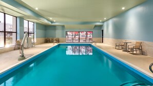 An indoor swimming pool with clear blue water, surrounded by beige walls and a light blue ceiling. There are chairs and tables on the right side, and a window on the left side.