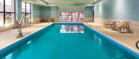 Una piscina interior con agua azul cristalina, rodeada de paredes beige y un techo azul claro. Se ven sillas y mesas a la derecha, y una ventana a la izquierda.