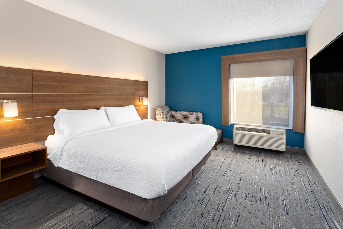 A modern hotel room with a large bed, bedside tables, a window with blinds, and a built-in air conditioning unit.