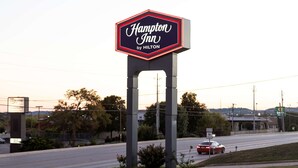 Ein Schild des Hampton Inn mit sechseckiger Form und rotem Rahmen, neben einer Straße, auf der ein Auto vorbeifährt.