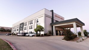 Ein Hampton Inn Hotel mit moderner Fassade, überdachter Zufahrt und einem Parkplatz mit mehreren Autos.