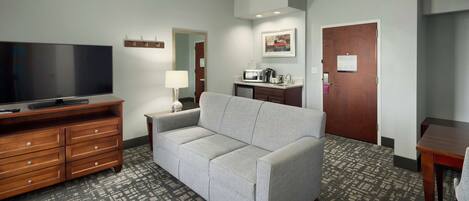 A hotel room with a gray sofa, wooden dresser, flat-screen TV, microwave, and a small table.