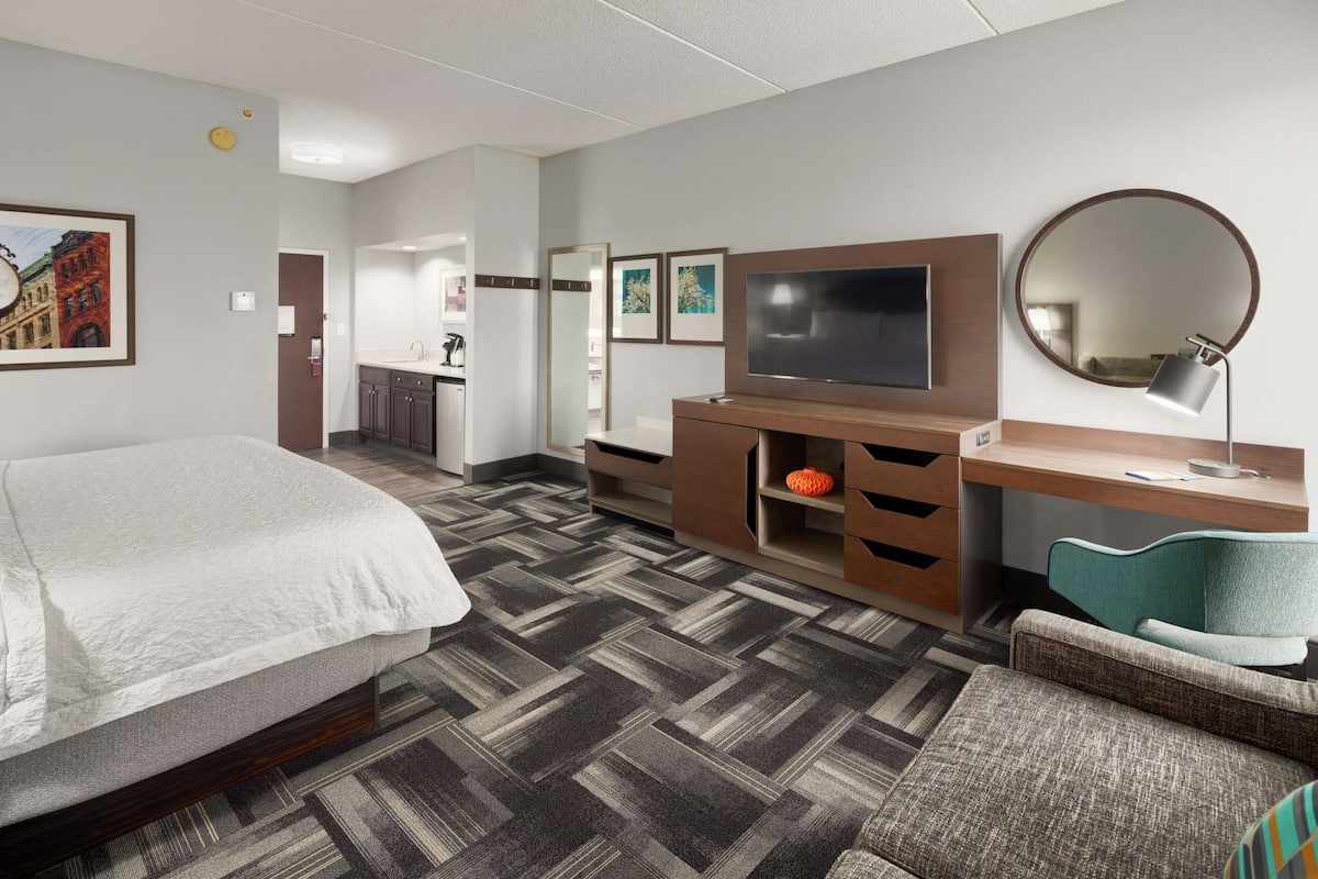 A hotel room with a bed, a TV, a desk with a chair, a mirror, and a bathroom visible through an open door.