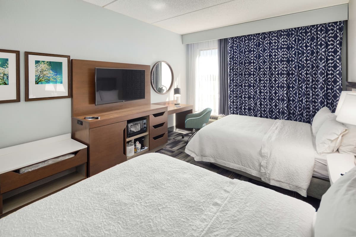 A hotel room with two beds, a TV, a desk with a chair, a mirror, and artwork on the wall.