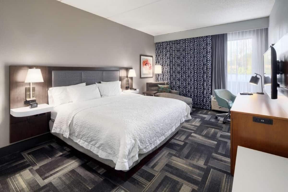 A hotel room with a large bed, bedside tables with lamps, a desk, a chair, and a TV.