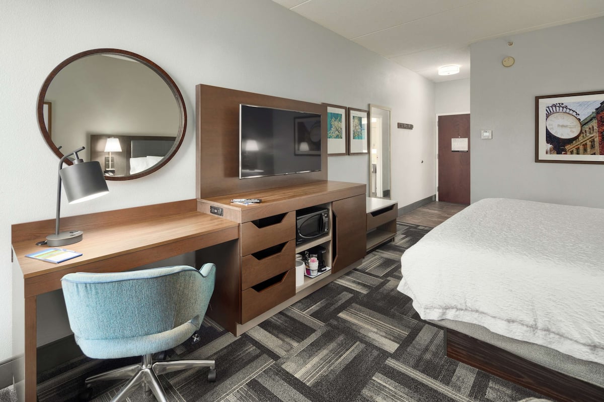 A hotel room with a wooden desk, a blue chair, a bed, a TV, a mirror, and artwork on the walls.
