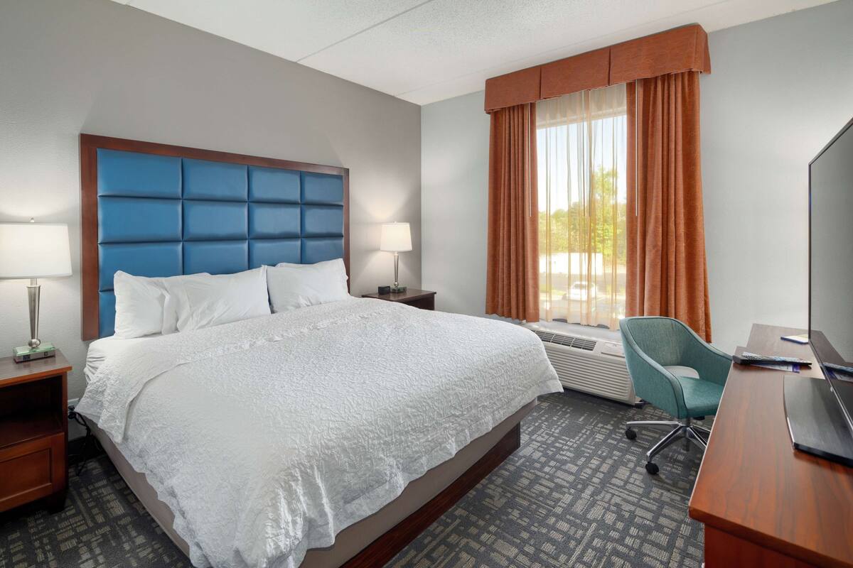A hotel room with a large bed, a desk, a chair, a TV, and a window with curtains.