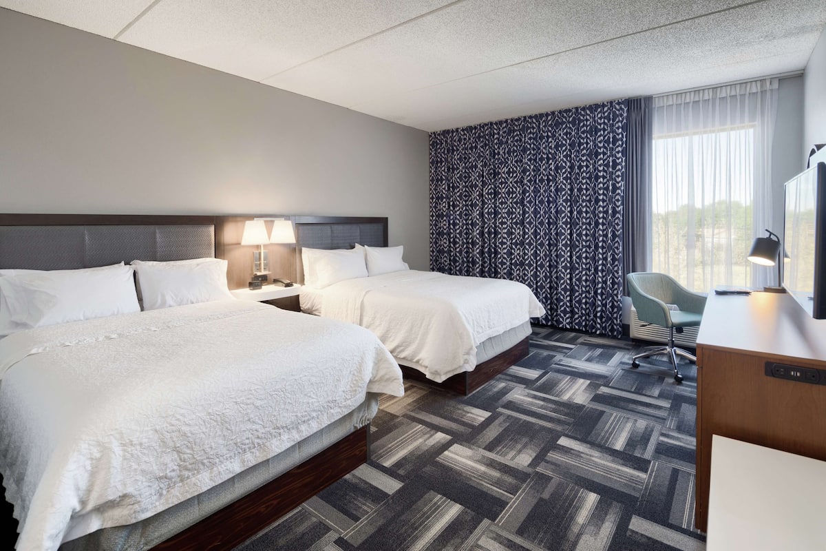 A hotel room with two beds, a desk, a chair, a patterned curtain, and a view of the outdoors.